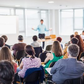 Speaker giving presentation on business conference. Business and entrepreneurship symposium. Speaker giving a talk at business meeting. Audience in conference hall. Rear view of unrecognized participant in audience.